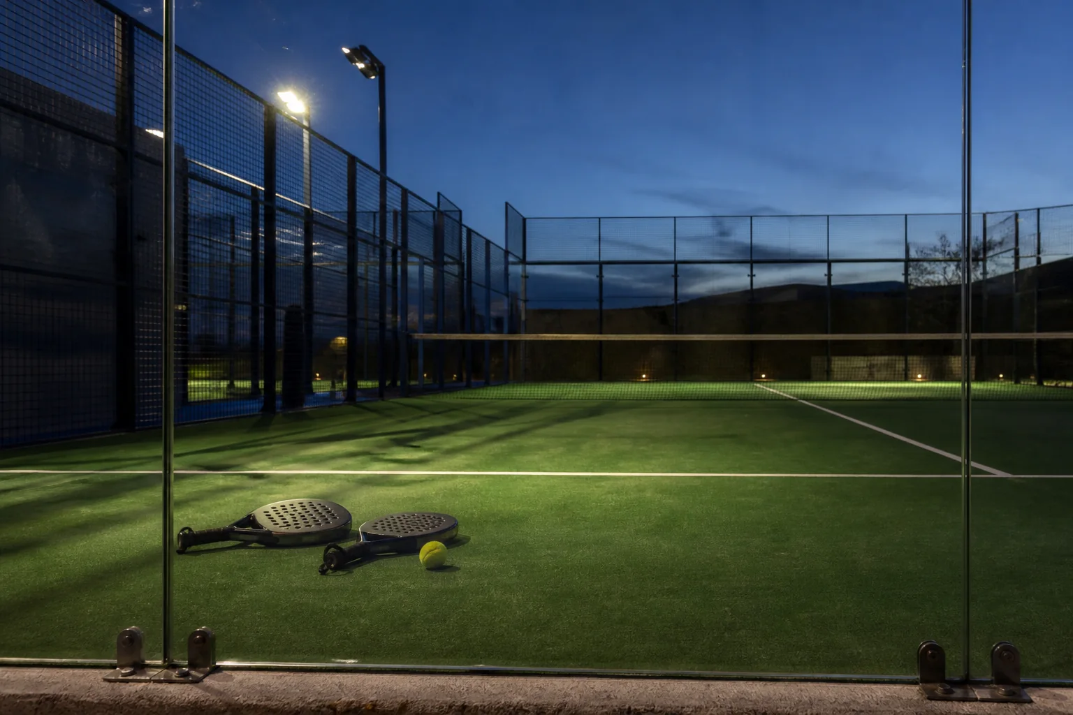 A padel court seen from behind the glass wall at dusk, two rackets and a ball resting on the service line